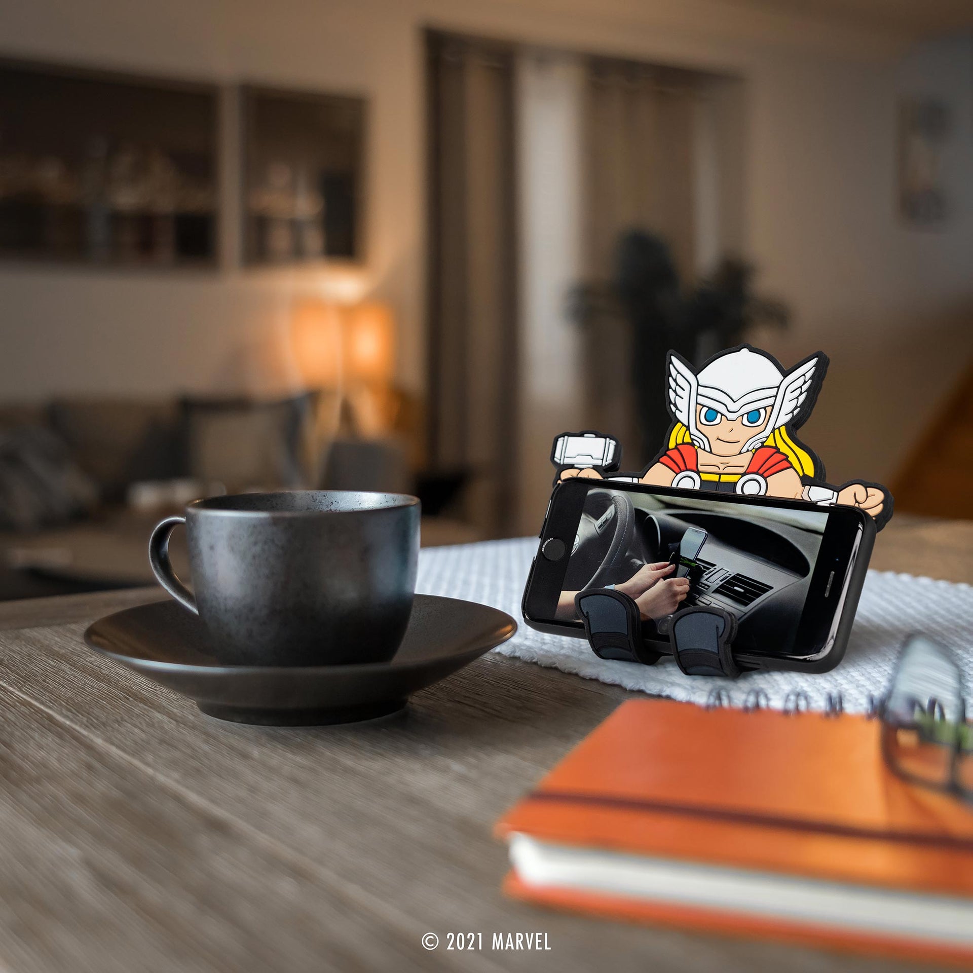 Image of Marvel Comics Thor Hug Buddy holding a cell phone on top of a kitchen table beside a warm cup of coffee or tea