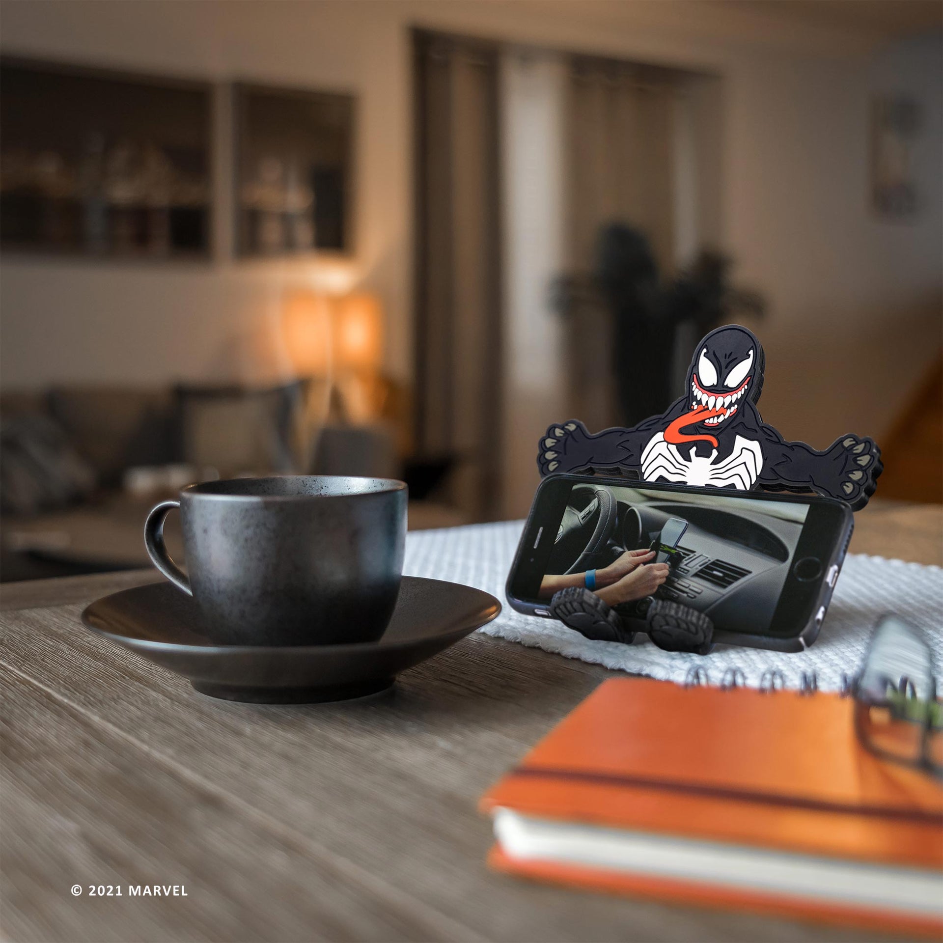 Image of Marvel Comics Venom Hug Buddy holding a phone displaying a video, on top of a kitchen table beside a warm cup of coffee or tea