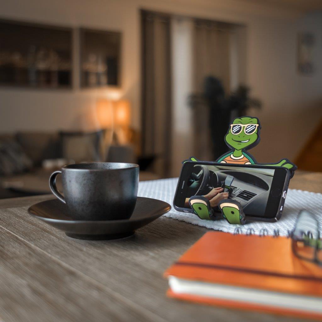 Image of Shellebrity the Turtle Hug Buddy holding a cell-phone sideways while a video plays on the screen, resting on its vent clip on top of a kitchen table, beside a book and a warm cup of coffee