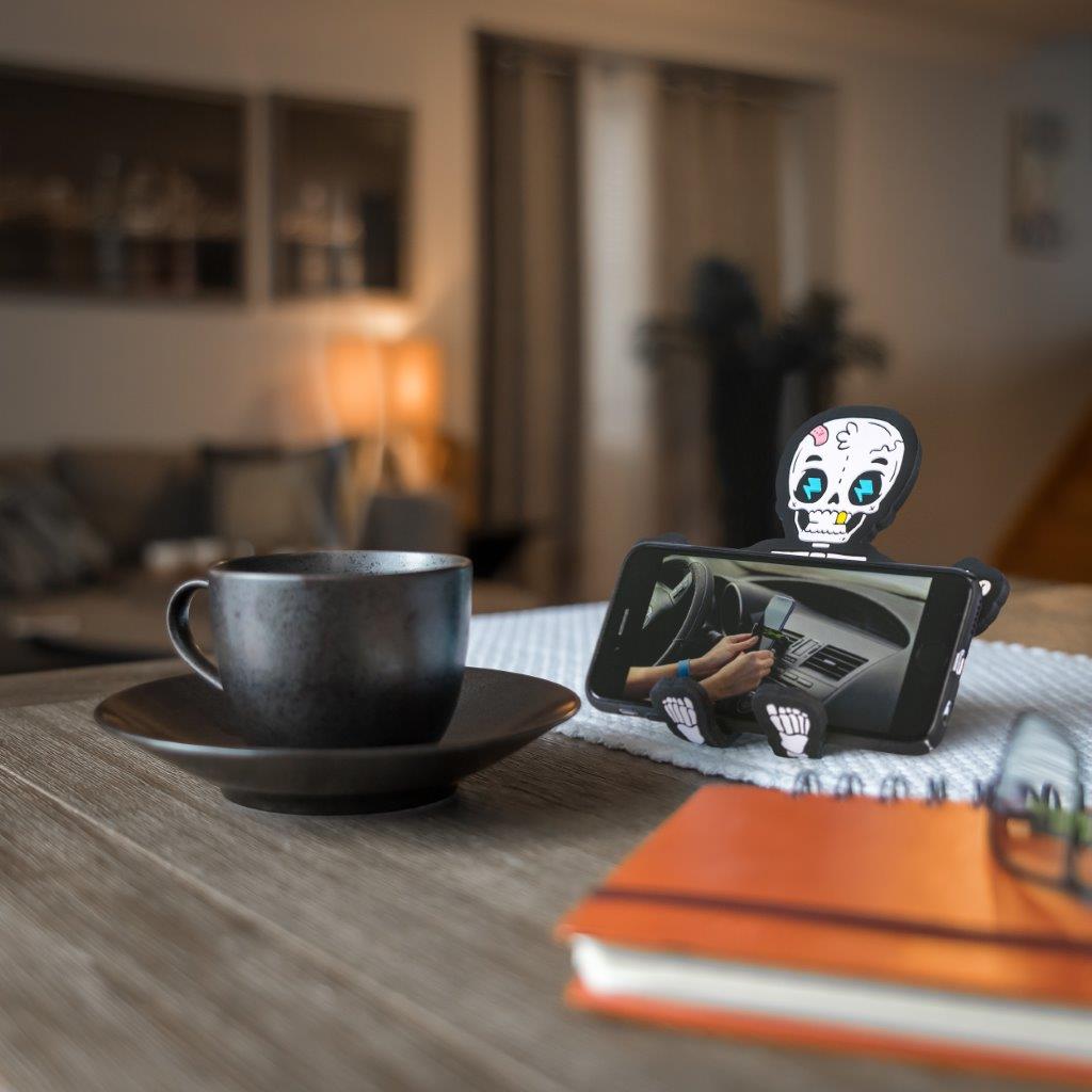 Image of Bones the Skeleton Hug Buddy holding a phone while sitting on a kitchen table beside a warm mug of coffee or tea