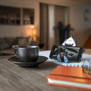 Image of Jaws the Shark Hug Buddy holding a cell phone while sitting on top of a kitchen table beside a warm cup of coffee or tea