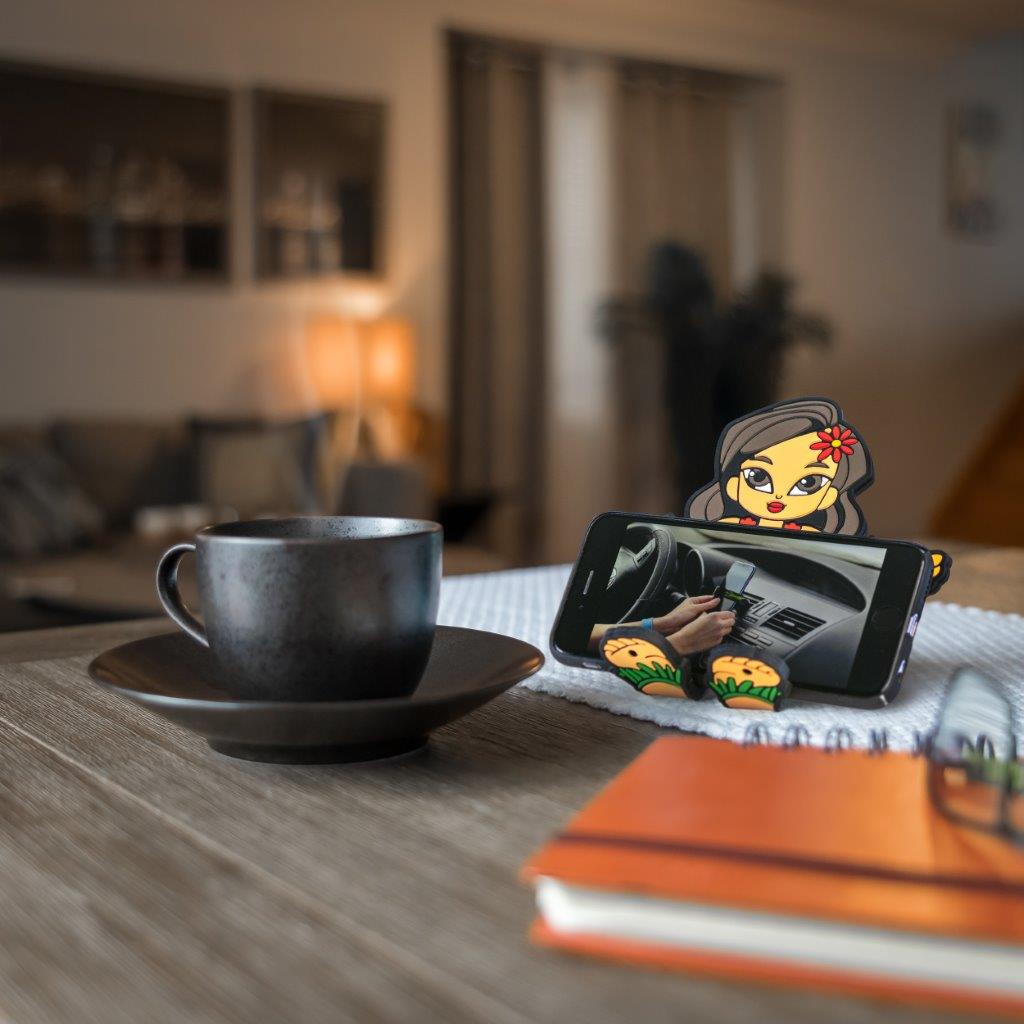 Image of Hula Girl Hug Buddy holding a cell phone while sitting on top of a kitchen table beside a warm cup of coffee or tea