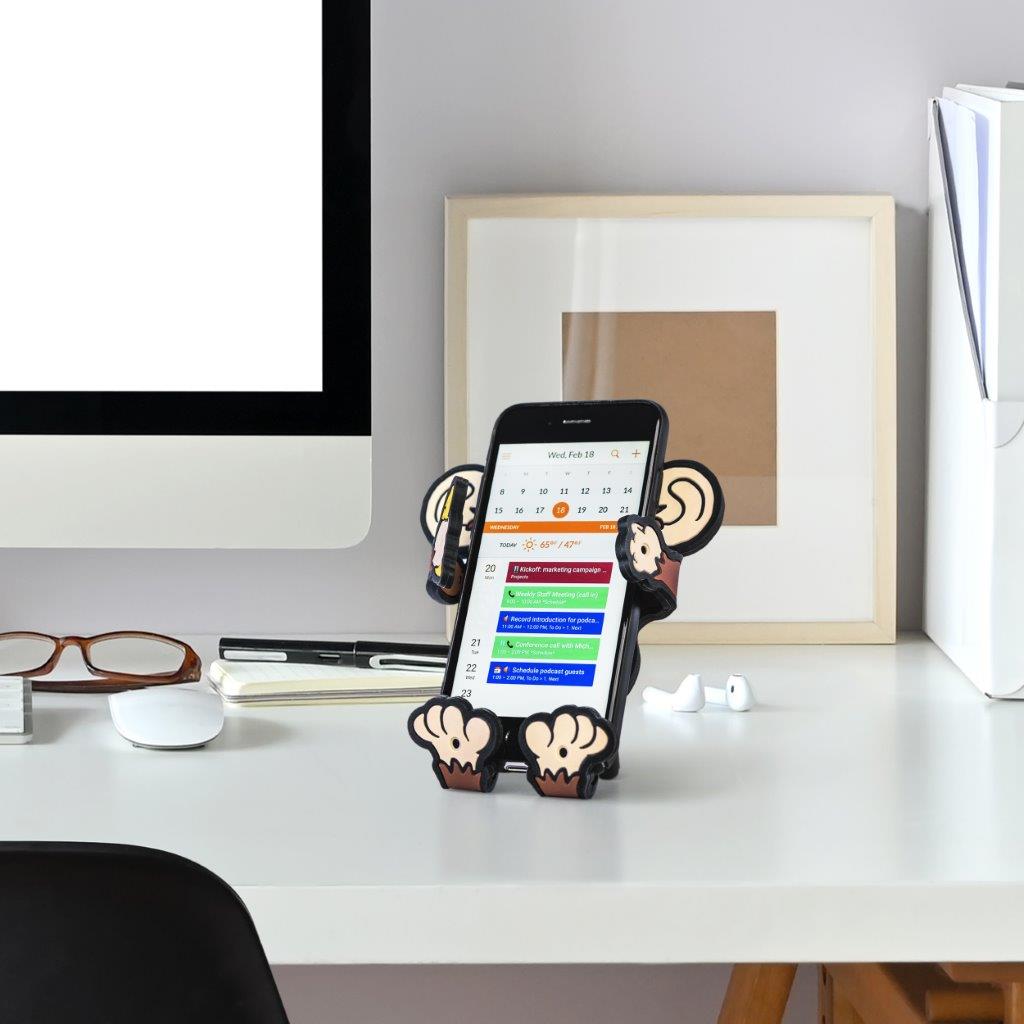 Image of the Bananas Monkey Hug Buddy sitting on a home office desk beside a computer screen holding a cell-phone, with the owners calendar open on the phone screen.