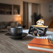 Image of Sloth Hug Buddy holding a cell phone on a kitchen table beside a warm mug of coffee or tea