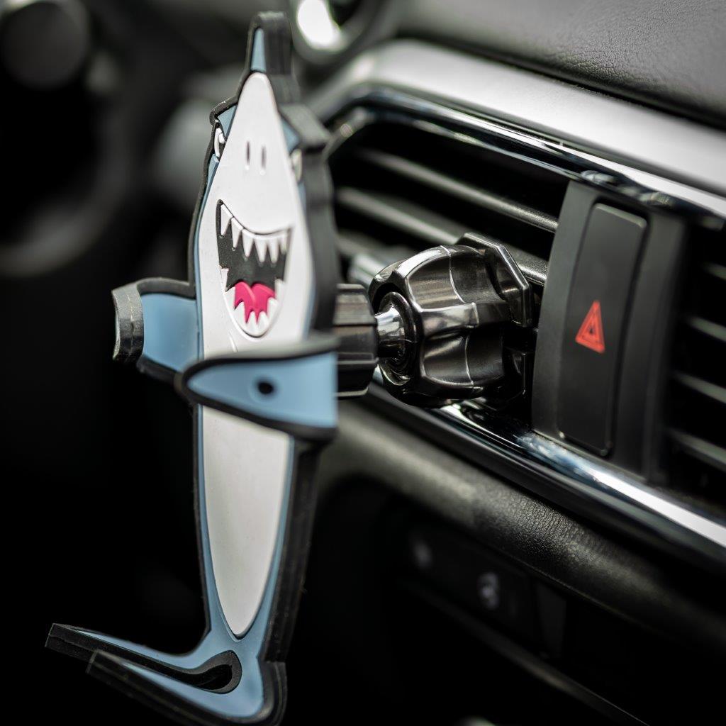 Image of Jaws the Shark Hug Buddy attached to a vehicle air vent with arms and legs folded in the closed position ready to grasp a phone or other smart device with a close up shot of the air vent clip