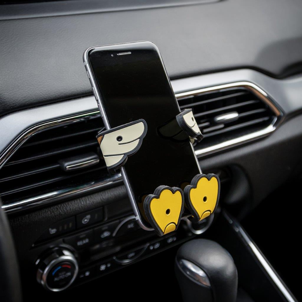 Image of Crumbs the duck Hug Buddy attached to a vehicle air vent holding a cell phone