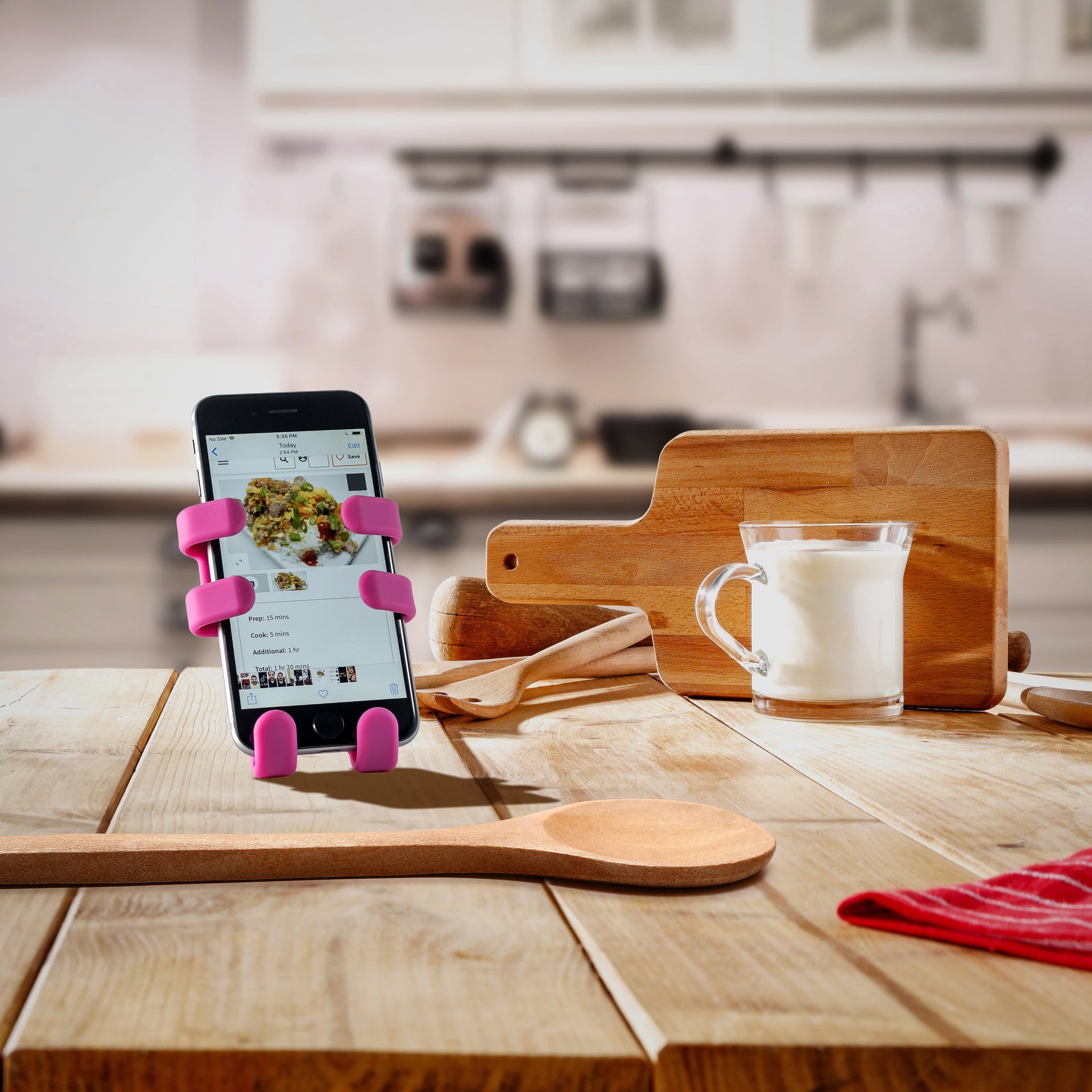 Image of OG Pink Hug Buddy holding a cell-phone with a recipe displayed on screen, on top of a kitchen counter surrounded by cooking utensils and ingredients