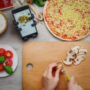 Image of OG Black Hug Buddy holding a cell-phone with a recipe displayed on screen, on top of a kitchen counter where you can see hands chopping mushrooms to place on a pizza