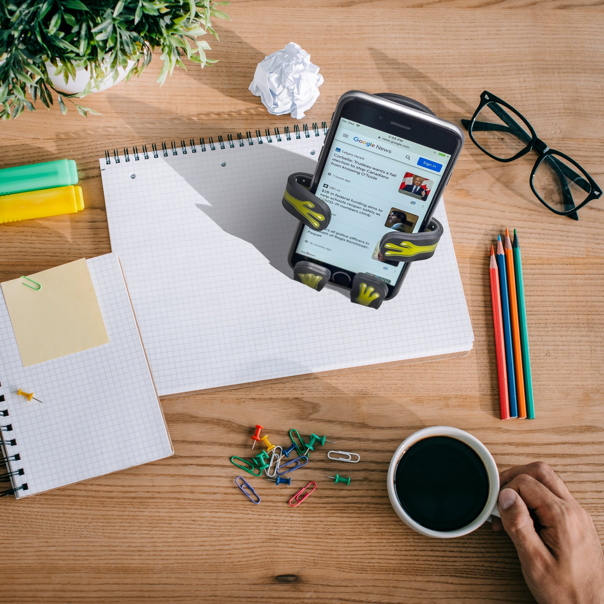 Image of Alien Hug Buddy sitting on a desk, holding a cell-phone with some news on the screen. A hand reaches in from off-screen holding a cup of coffee. The desk is full of paper and pencils, ready to tackle the tasks for your work day